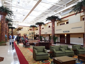 A hotel lobby with a high ceiling, palm trees, and a red carpet. There are seating areas and a reception desk.