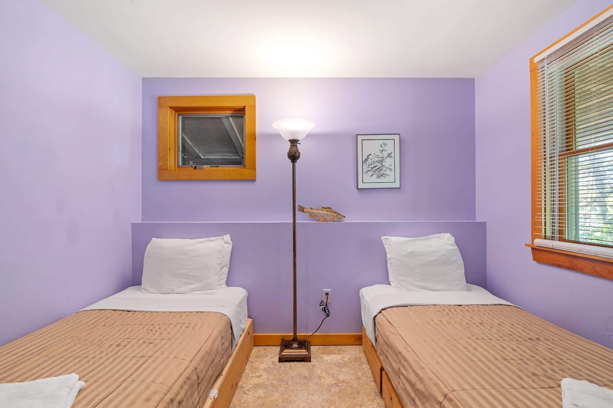 A room with two beds, a floor lamp, a window with blinds, and a framed picture on the wall.