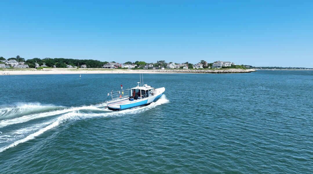 A boat on a calm body of water with a beach and houses in the background.