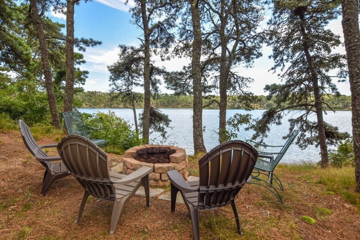 An outdoor seating area with two Adirondack chairs and a stone fire pit, surrounded by trees and overlooking a lake.