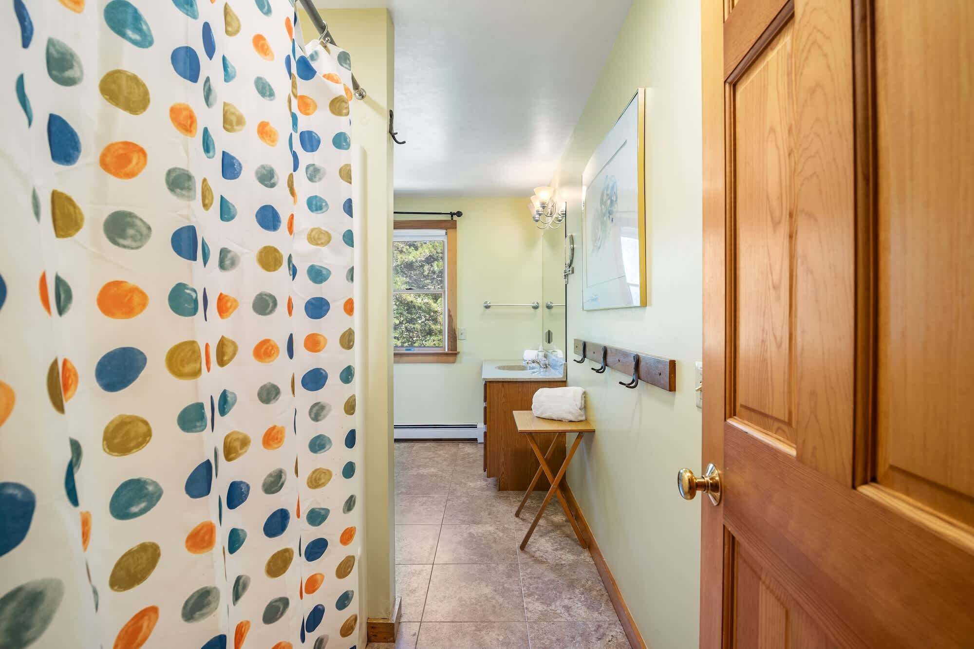 Un baño con cortina de ducha de lunares coloridos, una puerta de madera, un lavabo con una toalla y una ventana con vistas a la vegetación.