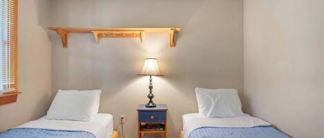 A small, cozy bedroom with two single beds, a nightstand with a lamp, and a wooden shelf above the beds.