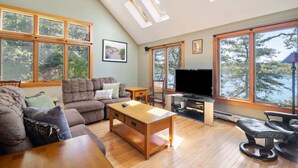 A cozy living room with a sofa, a coffee table, a TV, and a fireplace. There are large windows with a view of trees and a lake. - Amazing Location! Lakefront Waterside Deck (Chatham)
