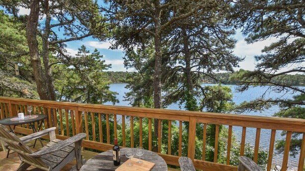 A wooden deck with a round table and two chairs, overlooking a lake with trees and a distant shoreline. - Amazing Location! Lakefront Waterside Deck (Chatham)