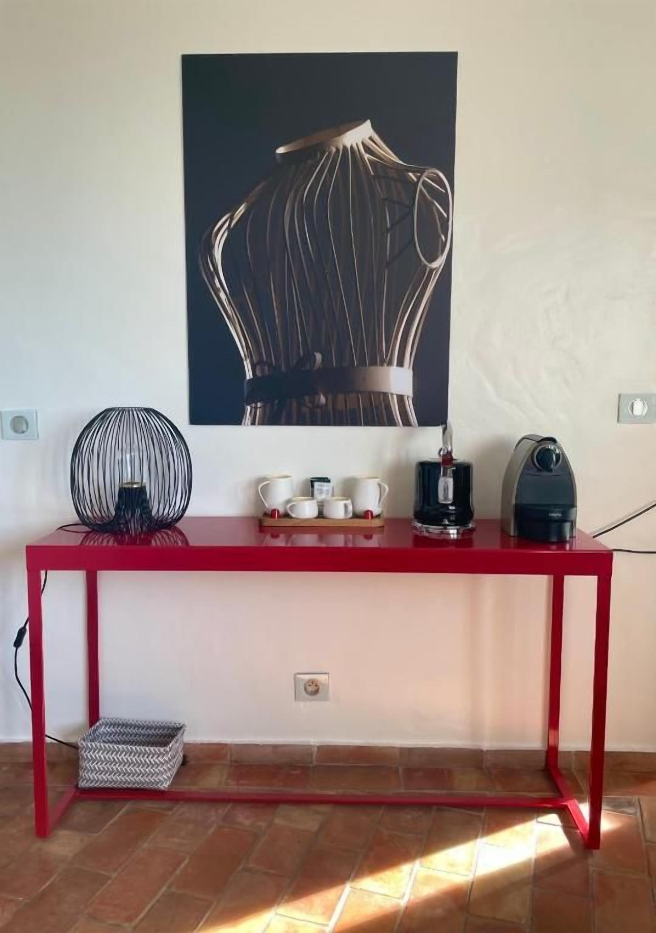 A red metal table with a black fan, white cups, and a coffee maker on it.