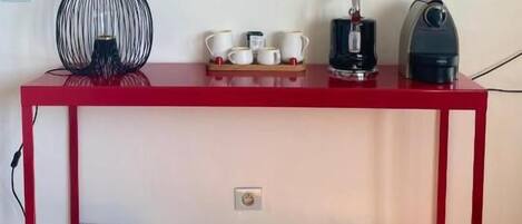 A red metal table with a black fan, white cups, and a coffee maker on it.