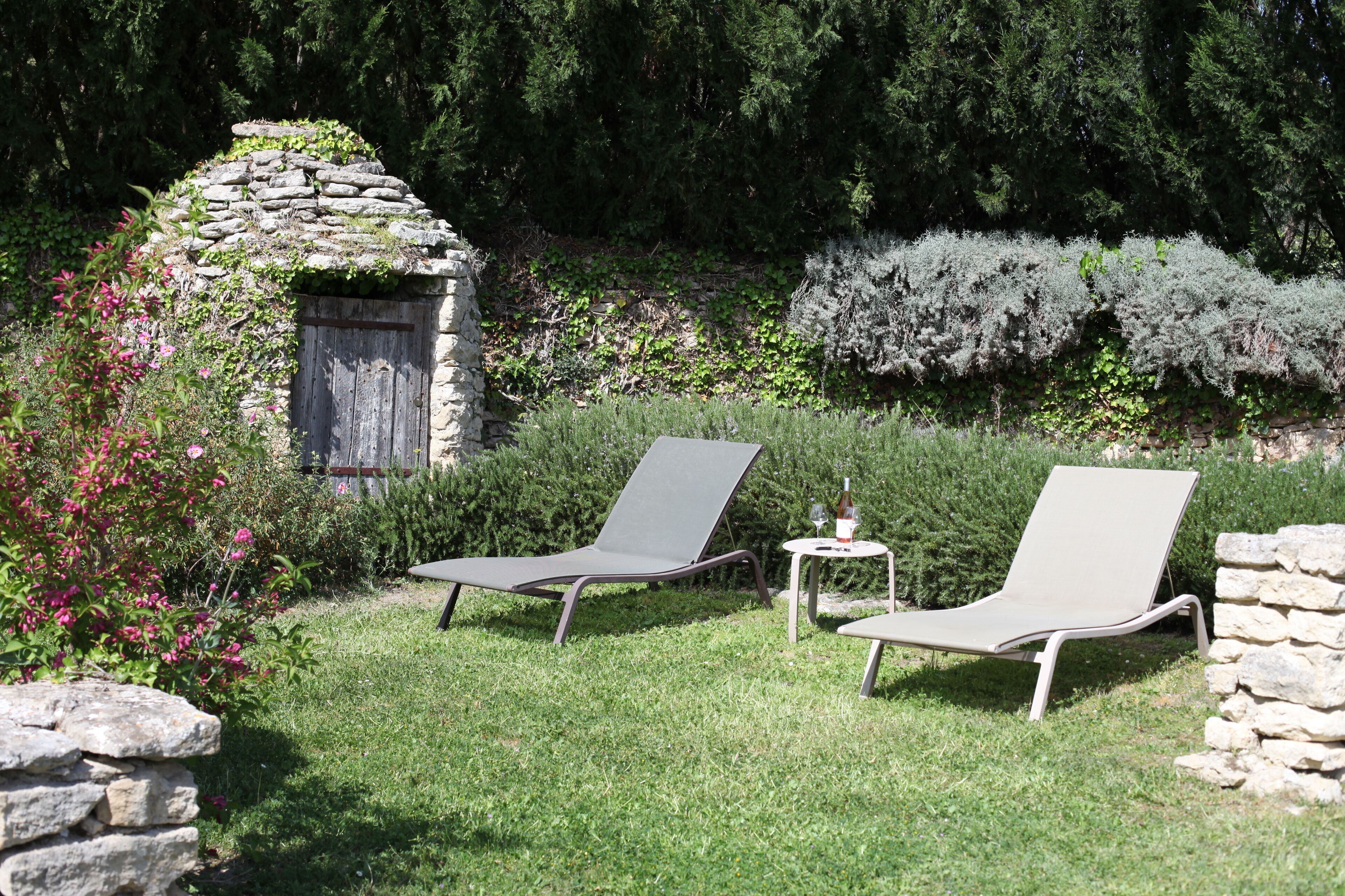 Two lounge chairs in a garden with a stone structure and lush greenery.