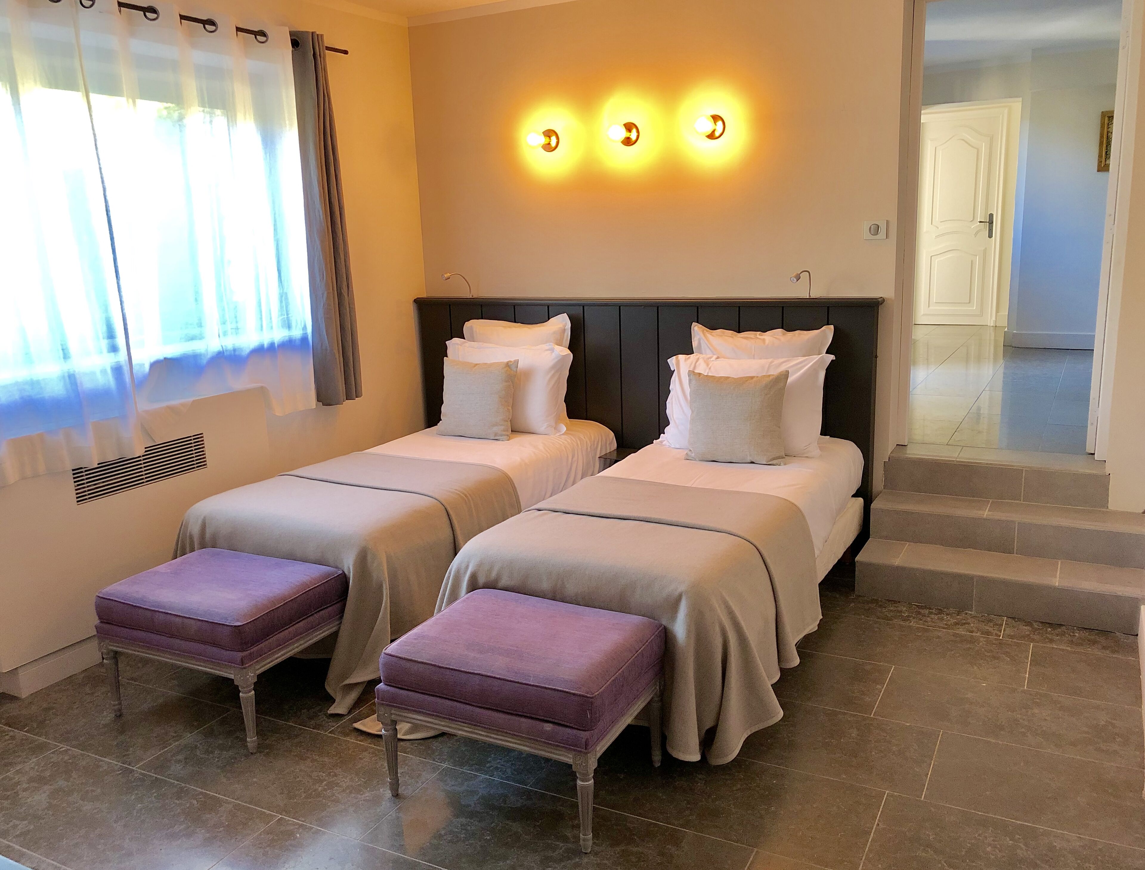 A hotel room with two beds, a window with curtains, a staircase, and a wall-mounted light fixture.