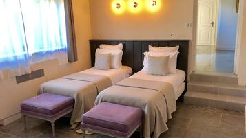 A hotel room with two beds, a window with curtains, a staircase, and a wall-mounted light fixture.