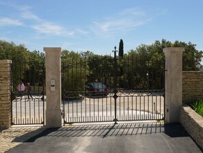 A gated entrance with a stone wall and metal gate, leading to a parking area with a car. - Domaine de l'Enclos (Gordes)
