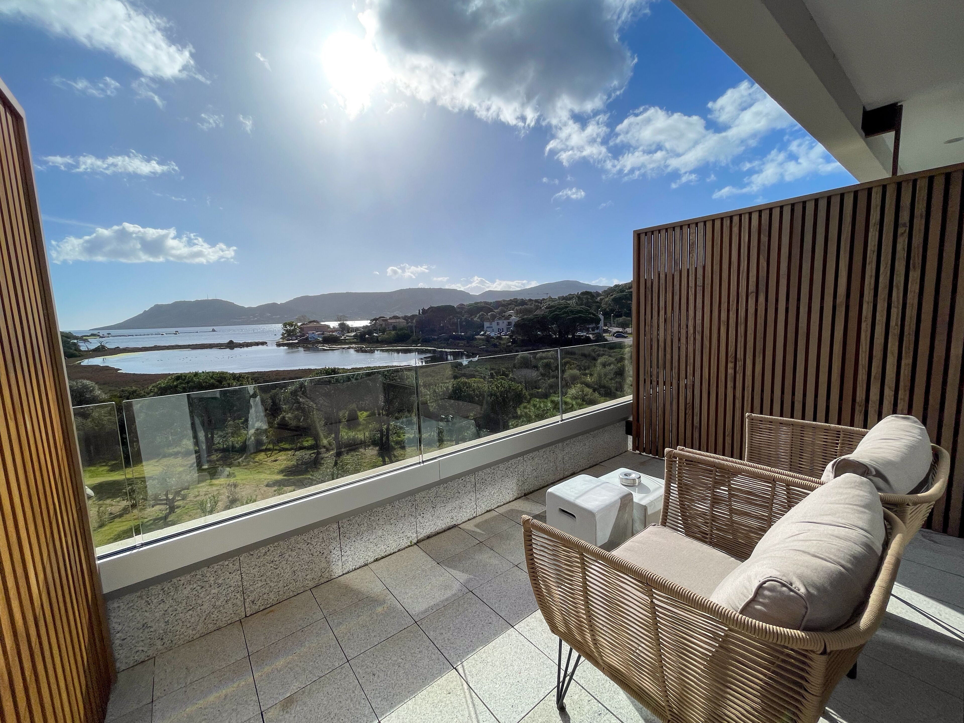A balcony with wicker furniture, a clear view of a coastal landscape, and a mountain in the distance.