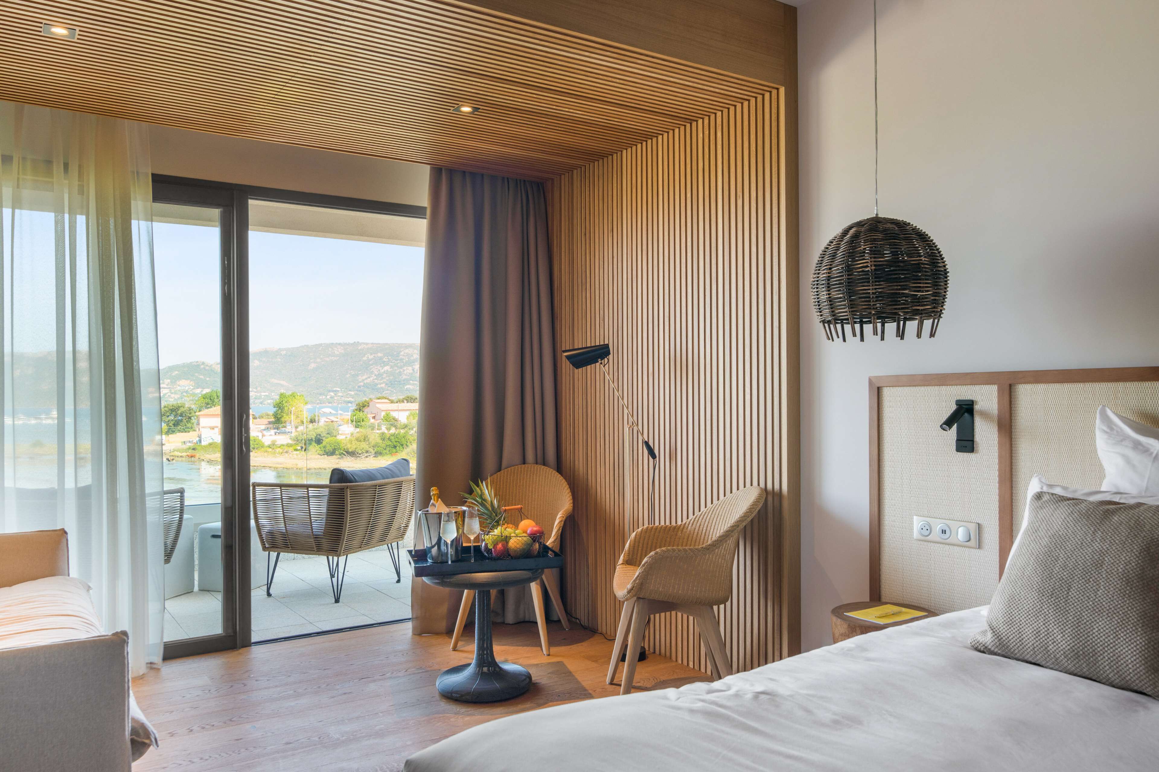 Quarto de hotel moderno com uma cama grande, uma mesa de jantar com frutas, duas cadeiras de vime e vista para o exterior, além de teto com ripas de madeira.