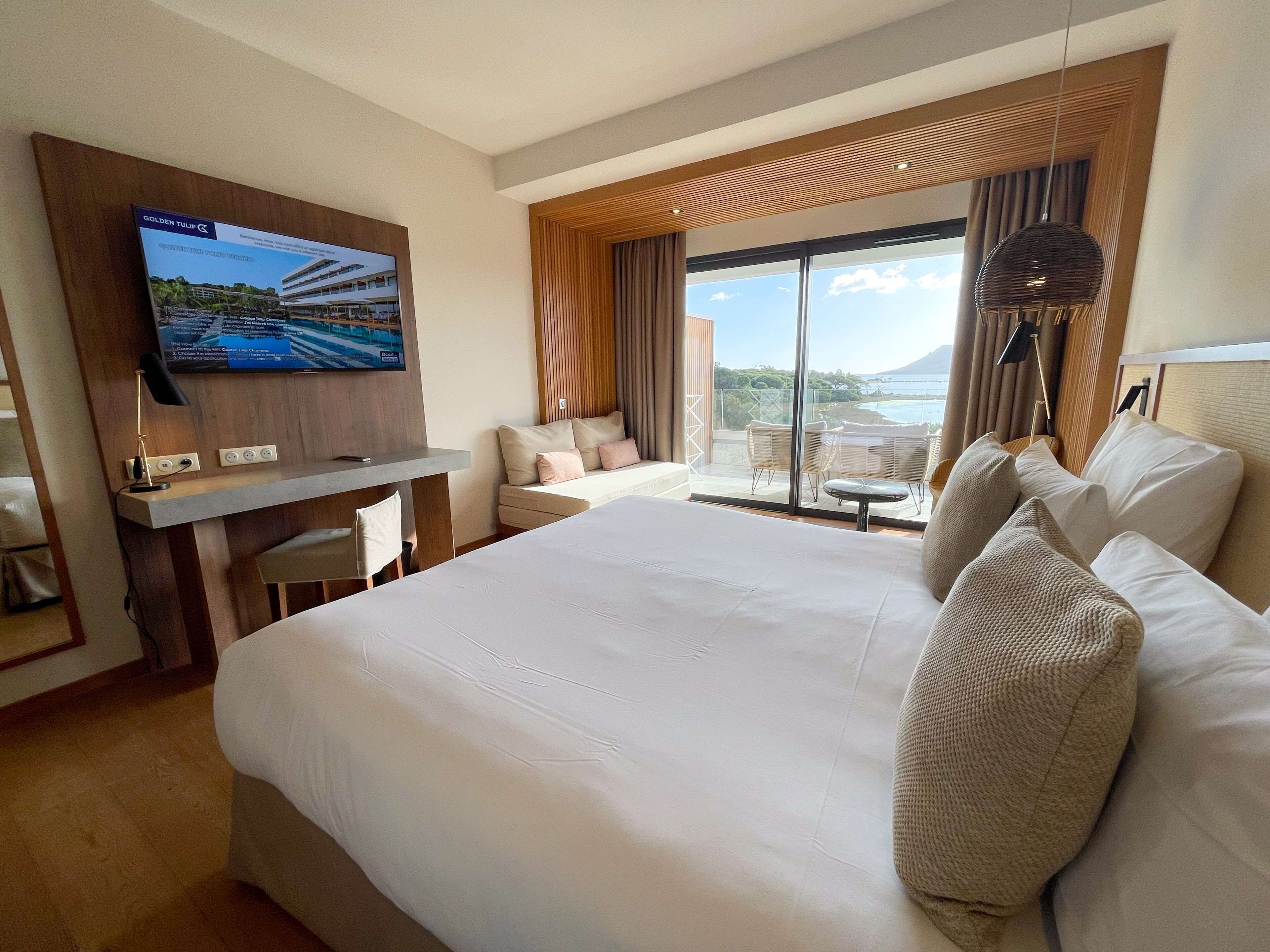 Ein Hotelzimmer mit einem großen Bett, einem Schreibtisch mit Fernseher, einem Balkon mit Meerblick und einem mit Vorhängen versehenen Fenster.