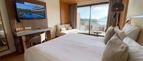Ein Hotelzimmer mit einem großen Bett, einem Schreibtisch mit Fernseher, einem Balkon mit Meerblick und einem mit Vorhängen versehenen Fenster.