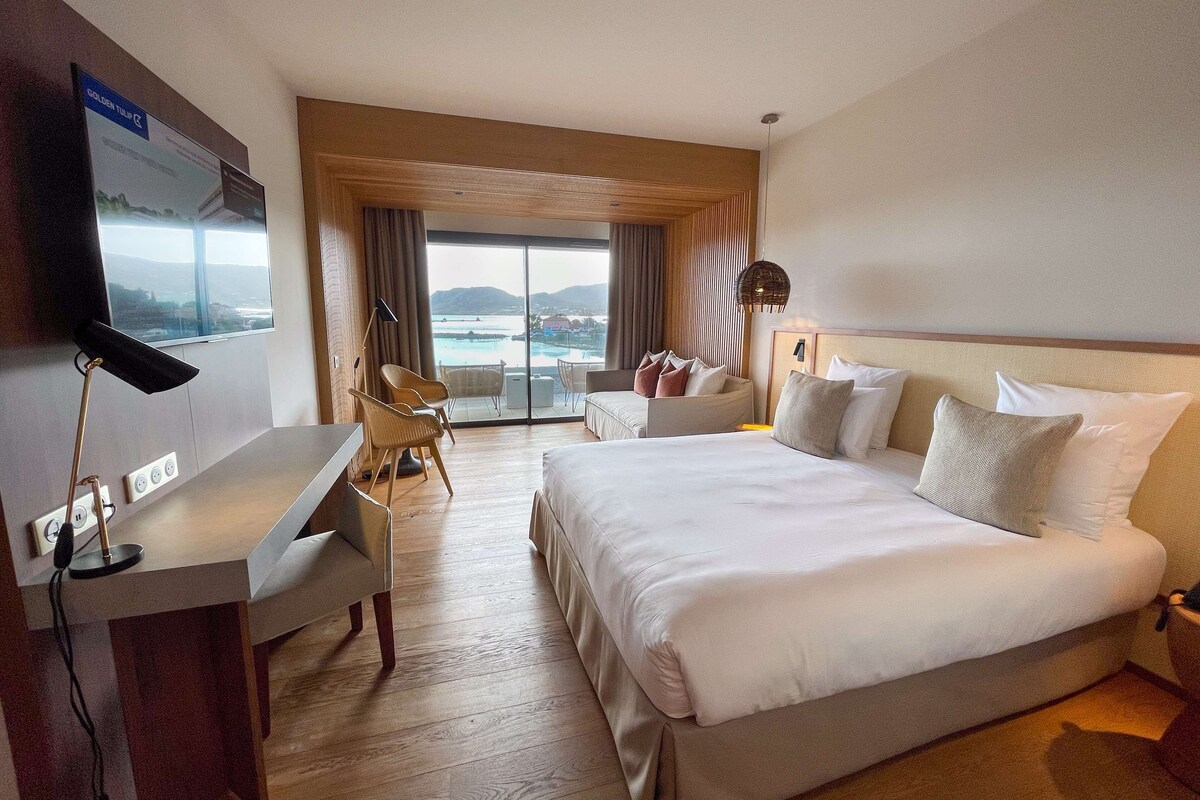A hotel room with a large bed, a desk, a chair, and a view of a body of water through a large window.