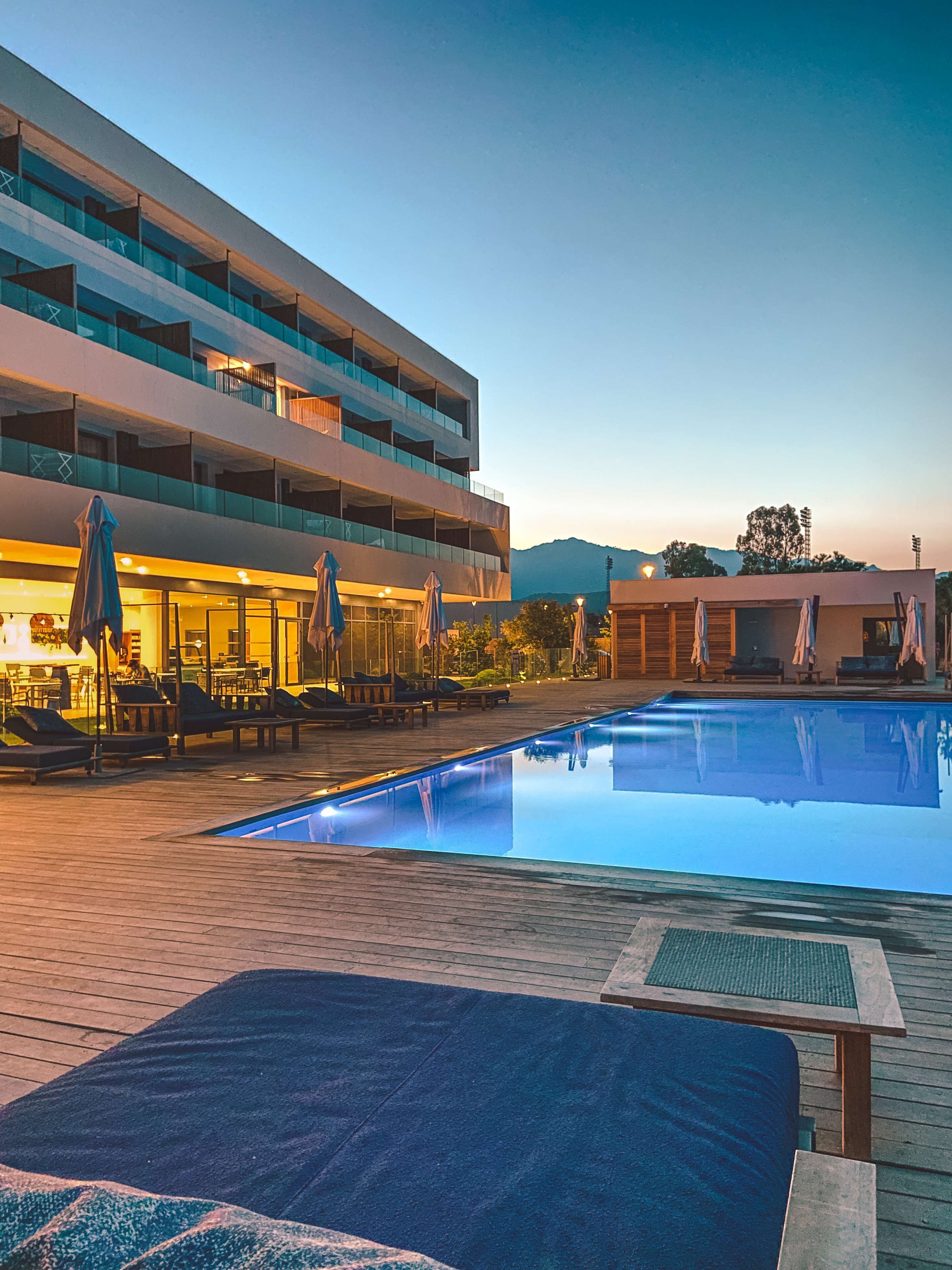 A swimming pool with lounge chairs and umbrellas, a building with multiple balconies, and a mountain in the background.