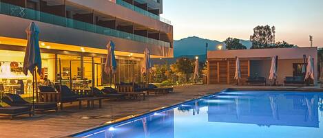 A swimming pool with lounge chairs and umbrellas, a building with multiple balconies, and a mountain in the background.