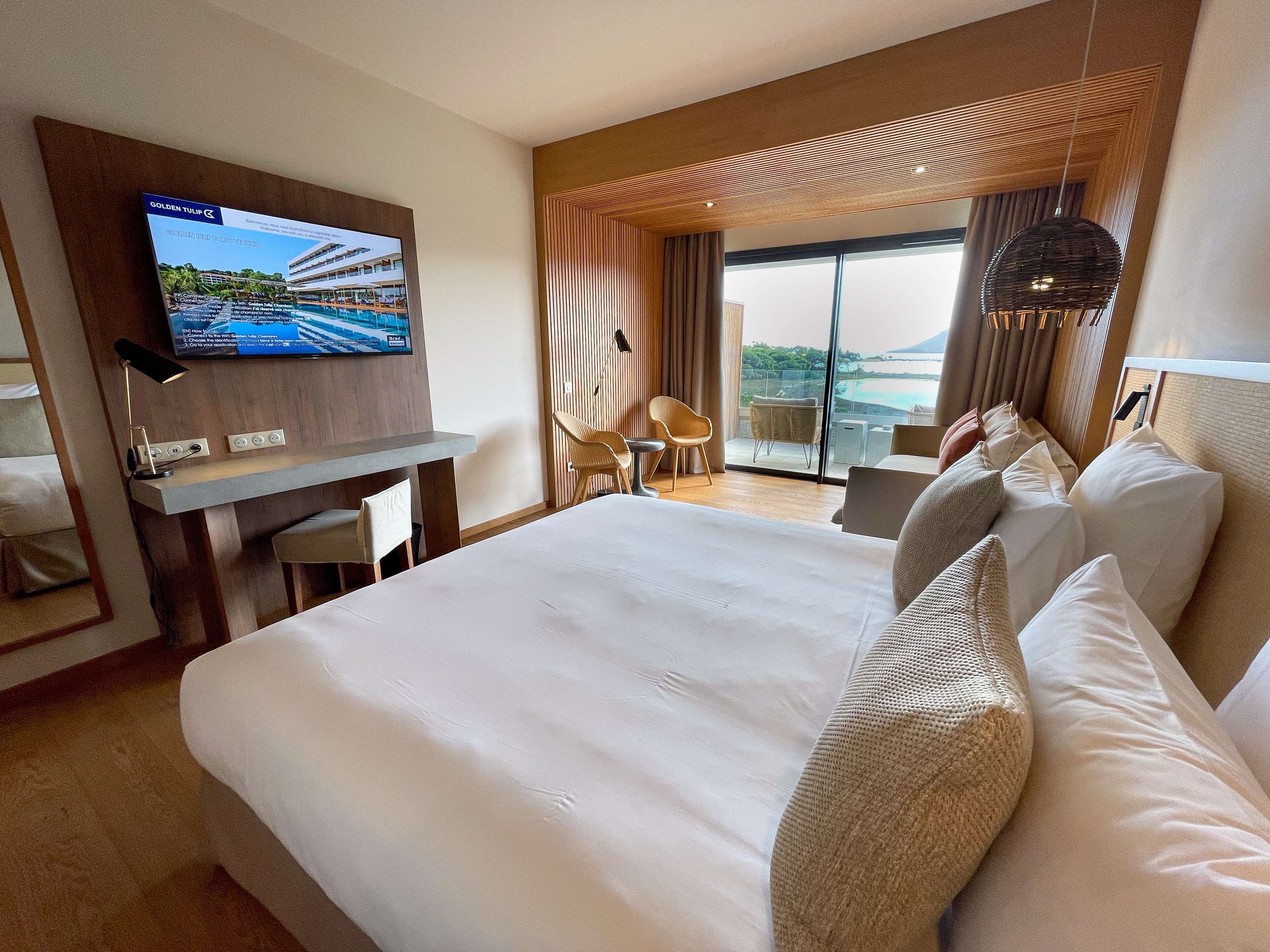 Ett hotellrum med en stor säng, ett skrivbord med en platt-TV, en balkong med utsikt och en modern inredning.