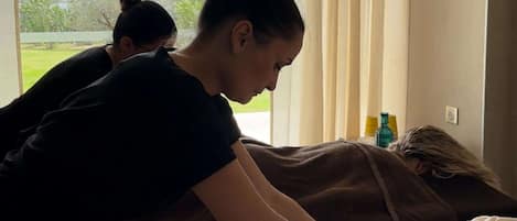 A massage therapy session in a room with a large window, a view of greenery outside, and a small table with bottles.