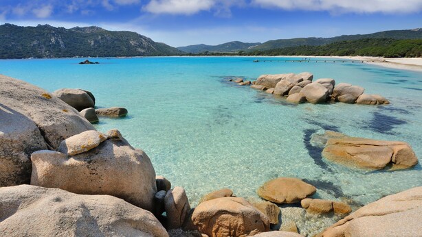 A clear turquoise sea with visible rocks in the foreground and a sandy beach in the background.