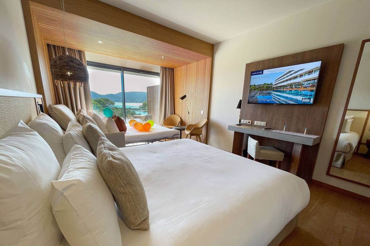 A hotel room with a large bed, a TV mounted on the wall, a desk with a chair, and a view of the outdoors.