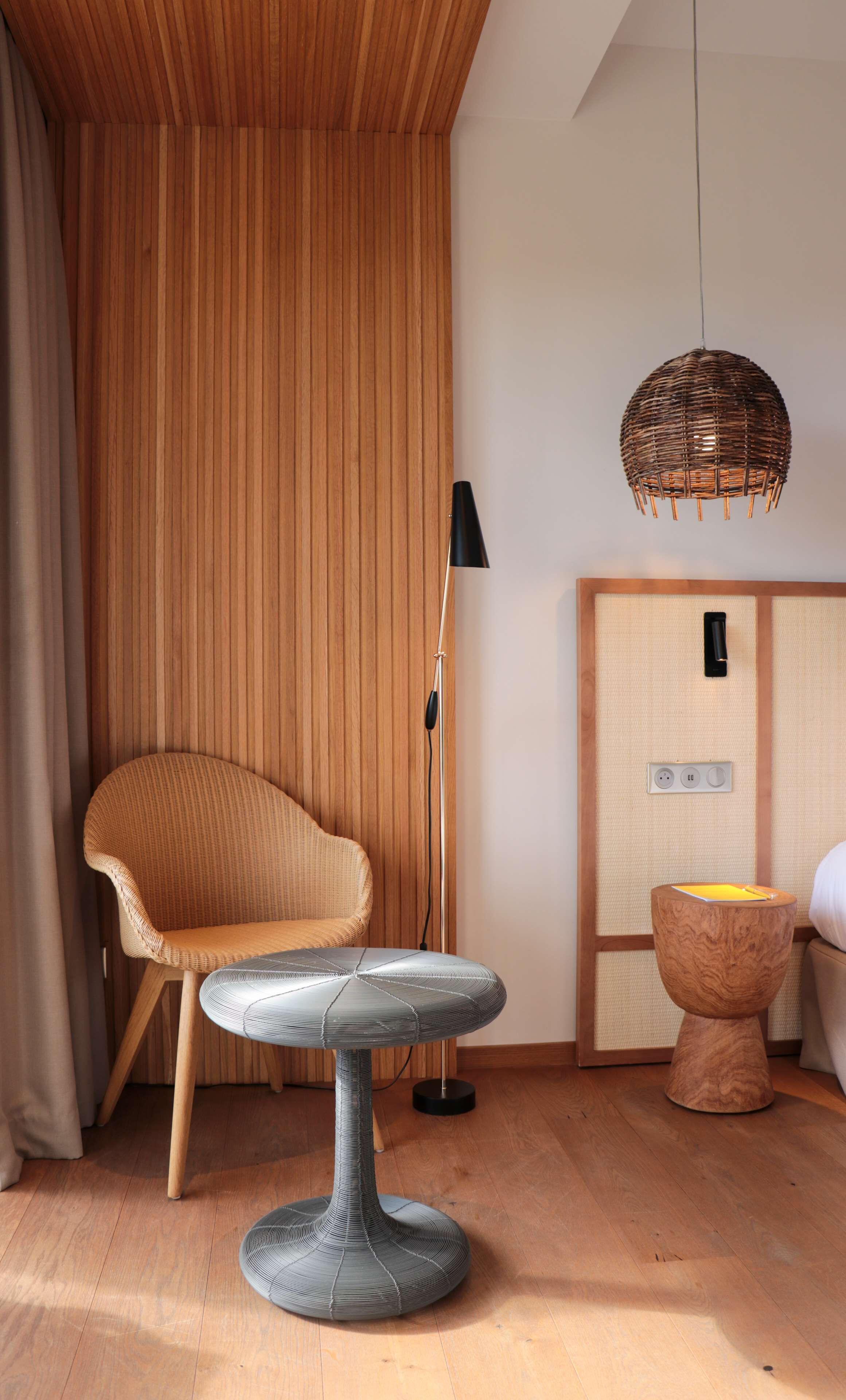 A room with a wooden slatted wall, a wicker chair, a modern table, a floor lamp, and a traditional Japanese cabinet.