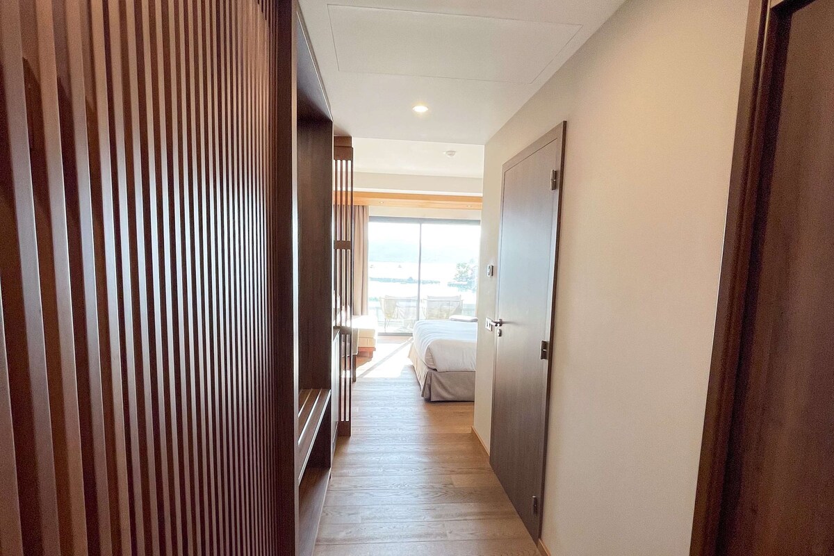 A hallway with wooden slats on one side, a bed with a white bedspread in the background, and a closed door on the right.