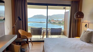 A hotel room with a large bed, a desk, two chairs, and a view of a body of water and hills.