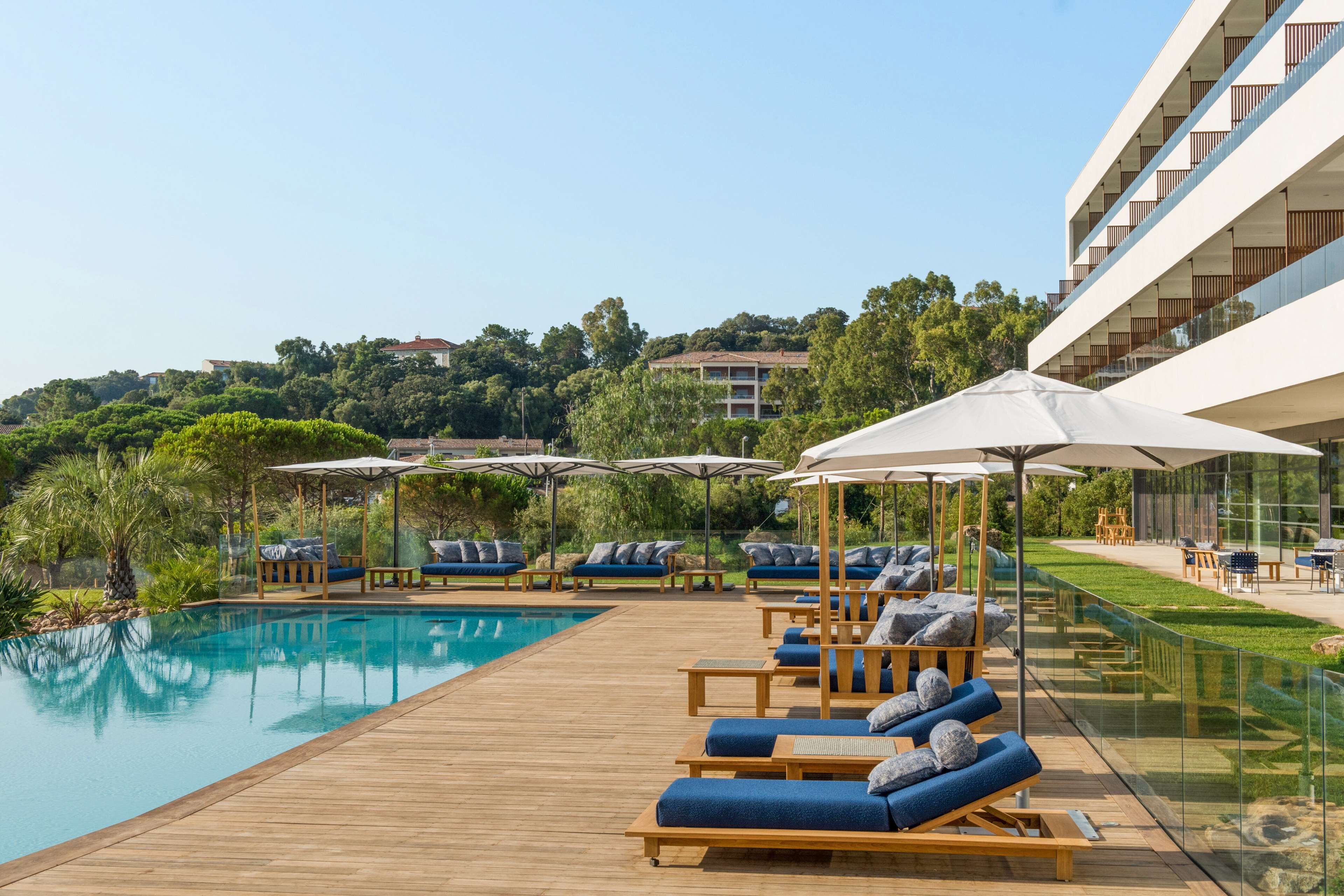 Ein Poolbereich mit Liegestühlen und Sonnenschirmen, ein Holzdeck sowie im Hintergrund ein Blick auf ein Gebäude und Bäume.