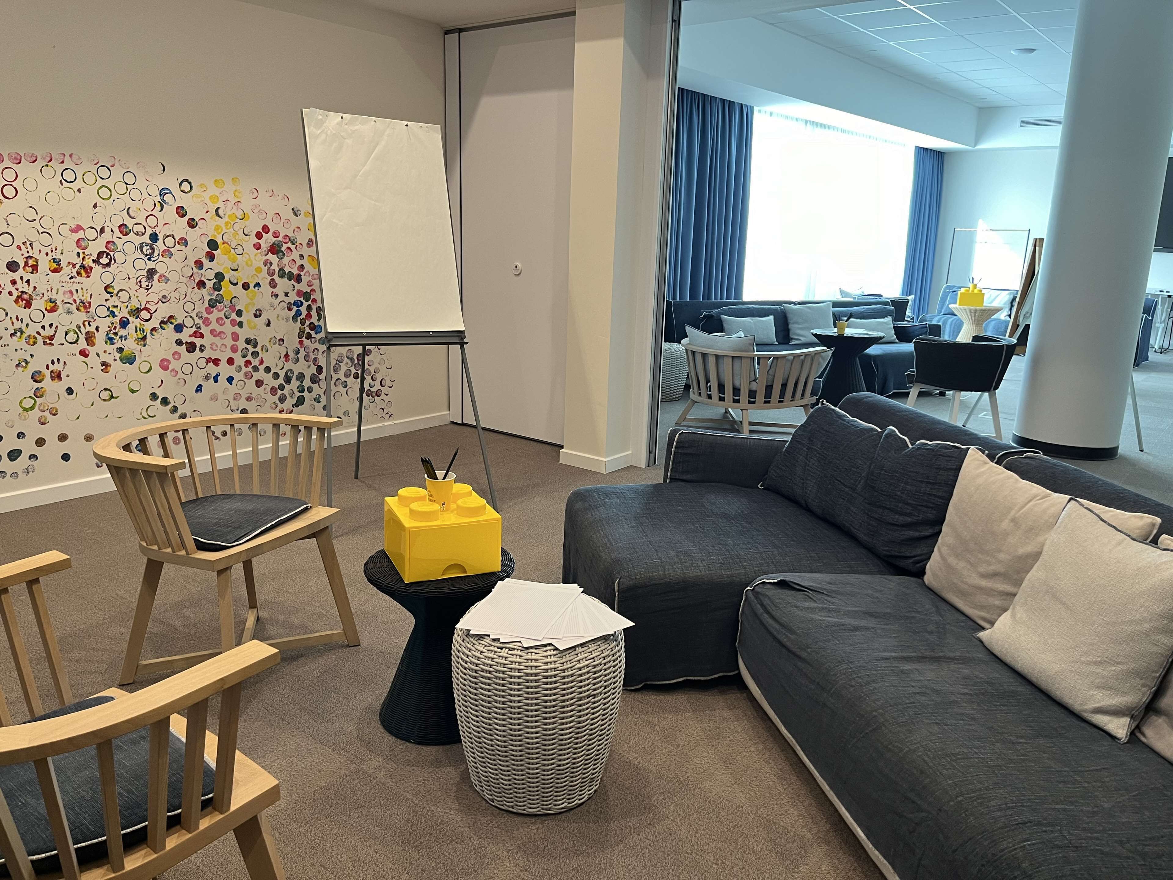 Ein modernes Wohnzimmer mit einem grauen Sofa, Holzstühlen, einem kleinen gelben Tisch und einem farbenfrohen Wandbild.
