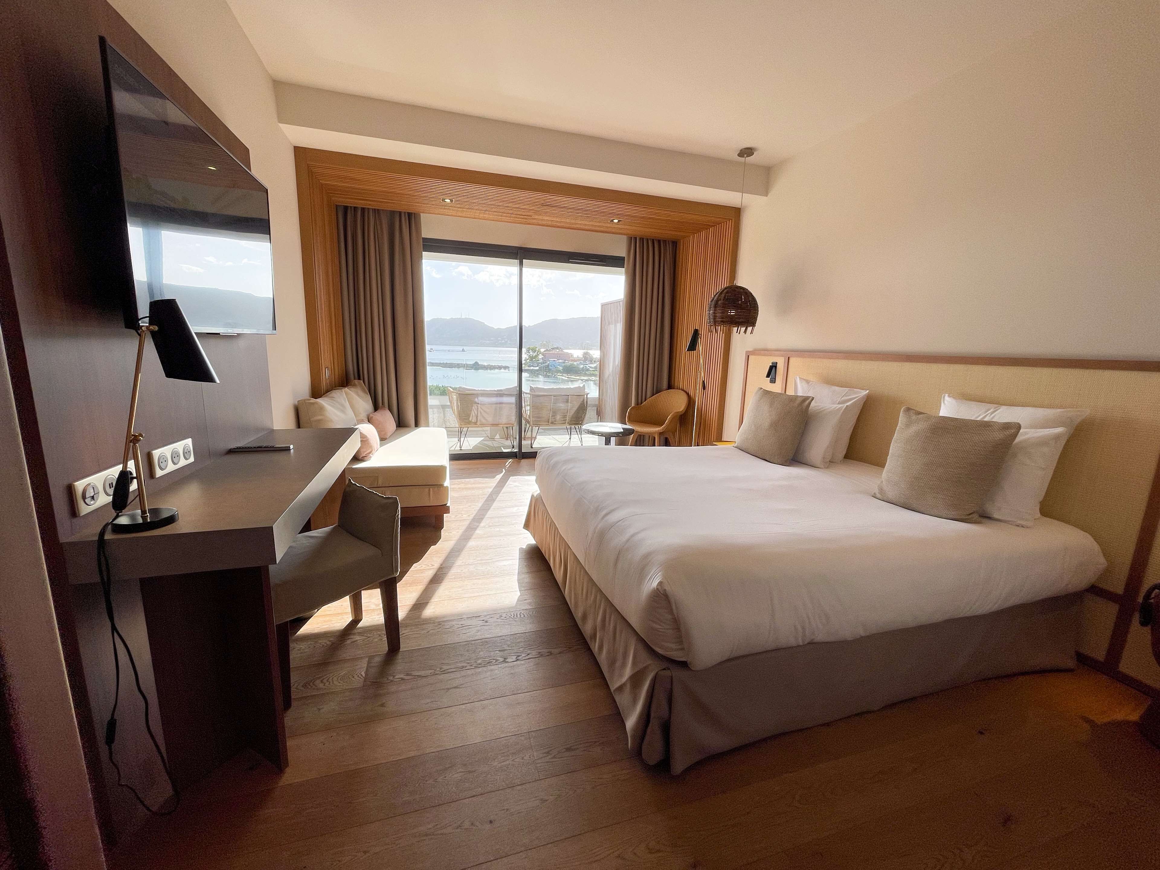 A hotel room with a bed, a desk, a chair, a TV, and a view of the outdoors.