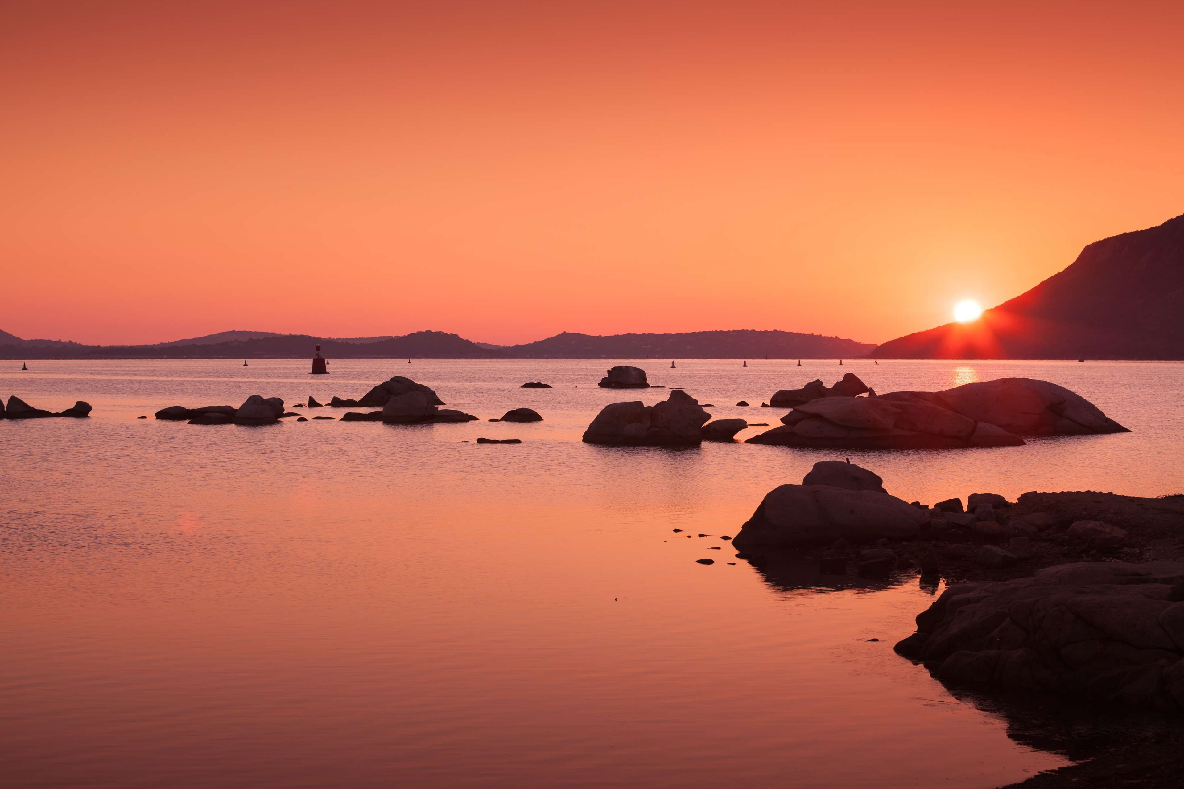Un coucher de soleil sur une étendue d’eau calme, avec des rochers et une colline au loin.
