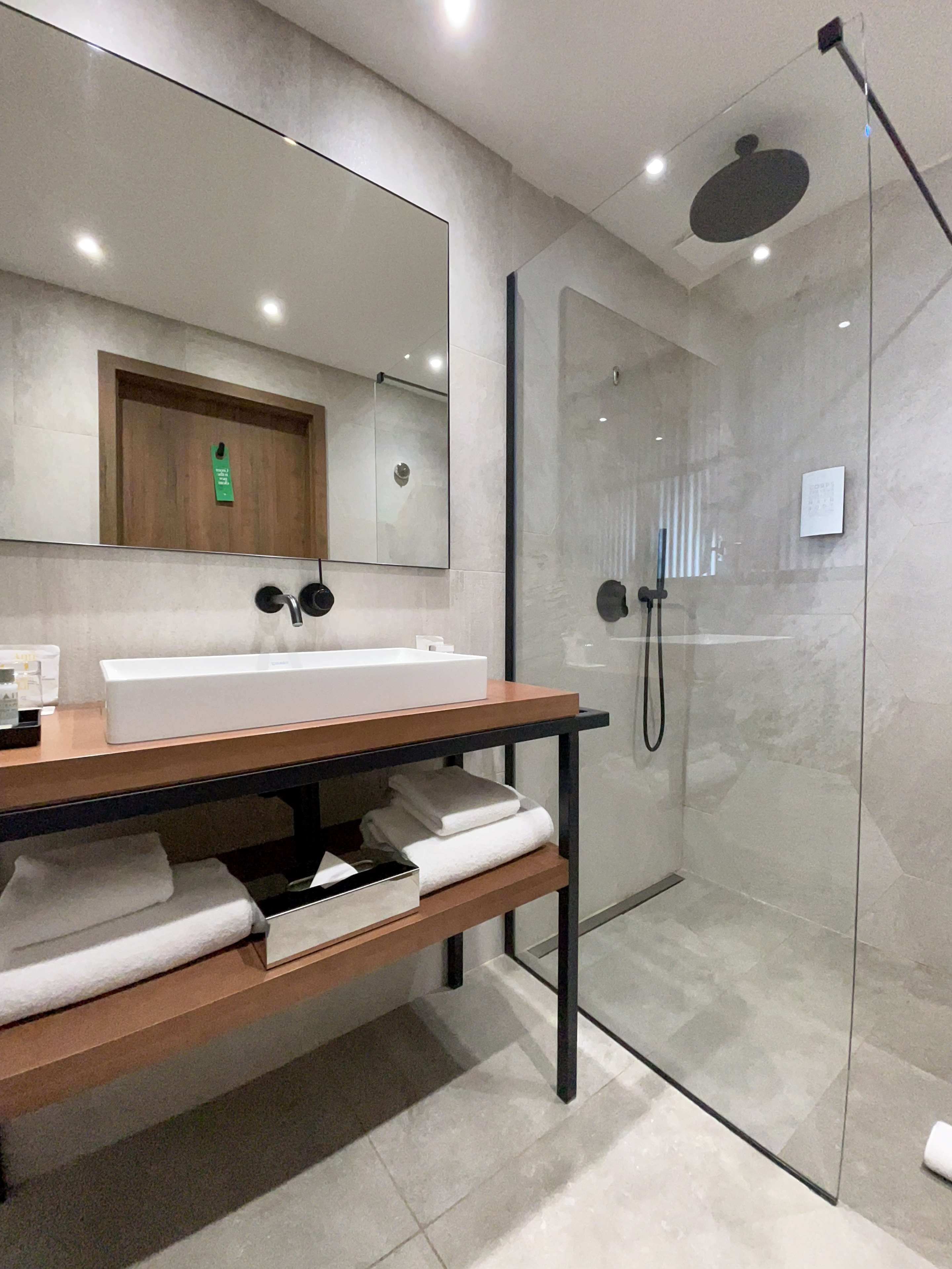 Un baño moderno con ducha envuelta en vidrio, tocador de madera con lavamanos blanco y un espejo grande sobre el tocador.