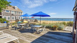 A wooden deck with outdoor furniture, including chairs and tables, overlooking a beach with boats in the water. - Beachfront with shared sundeck! (Provincetown)