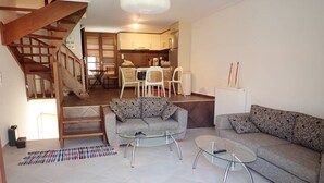 Uma sala de estar moderna com uma escada, dois sofás, uma mesa de centro de vidro e uma cozinha ao fundo.