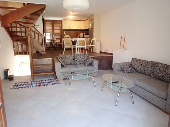 A modern living room with a staircase, two sofas, a glass coffee table, and a kitchen in the background.