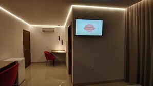 Moderni hotellihuone, jossa on seinälle asennettu taulutelevisio, punainen tuoli ja pieni työpöytä.