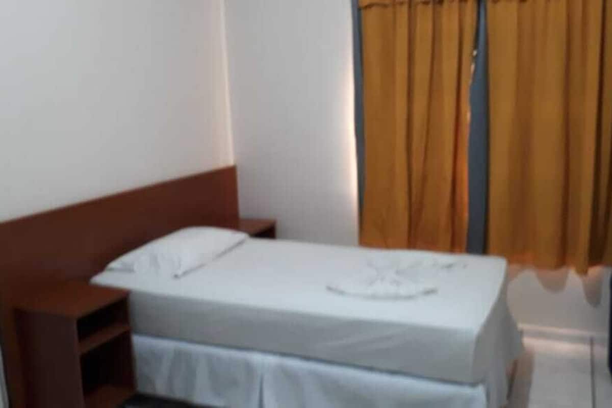 A single bed with a white mattress and a headboard, a nightstand, a blue chair, and a window with yellow curtains.