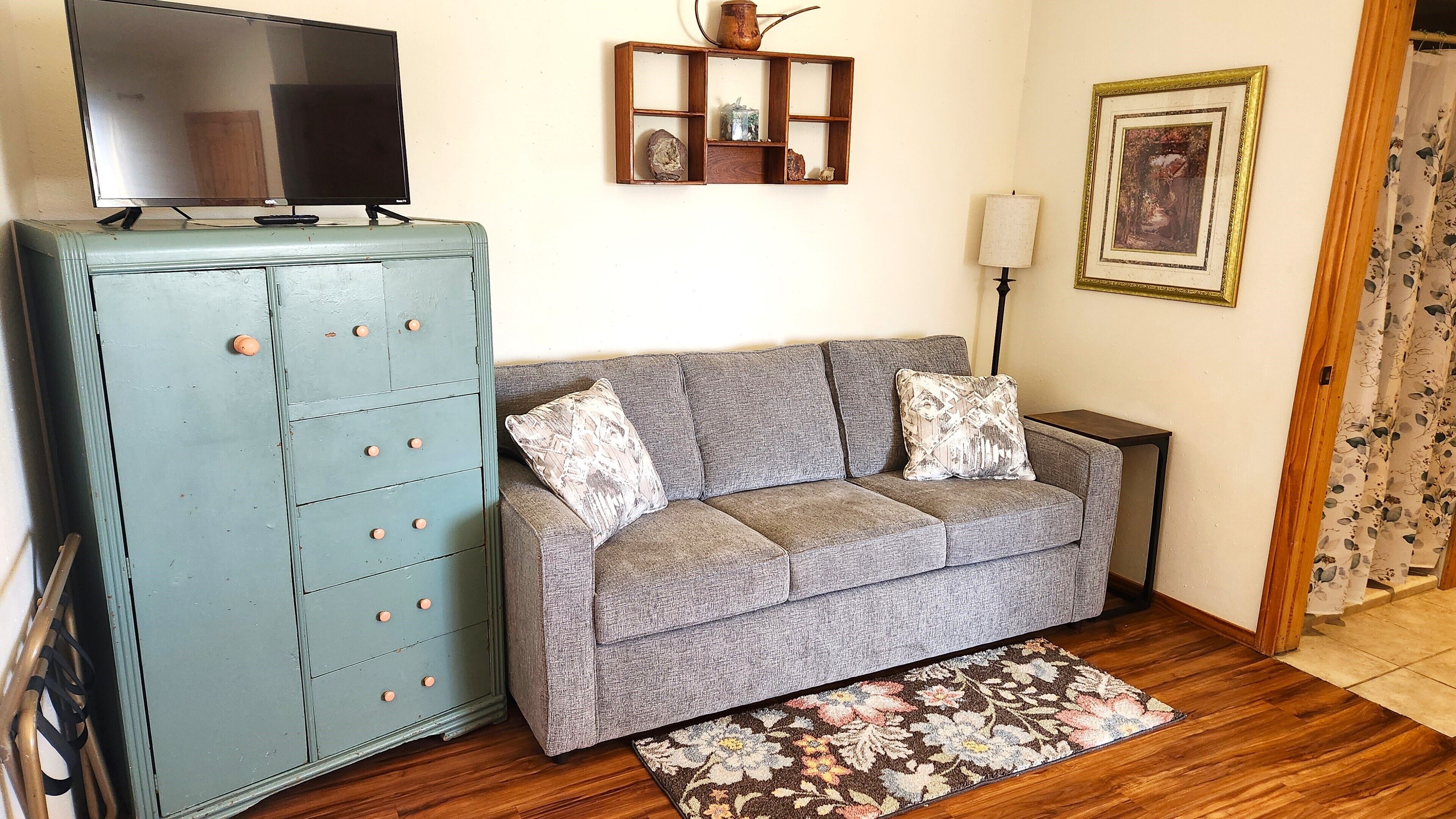 A living room with a grey sofa, a blue cabinet, a flat-screen TV, a decorative shelf, a lamp, and a framed picture on the wall.