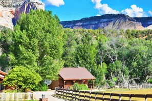 A rustic cabin with a wooden exterior and a red roof, surrounded by lush greenery and mountains in the background.