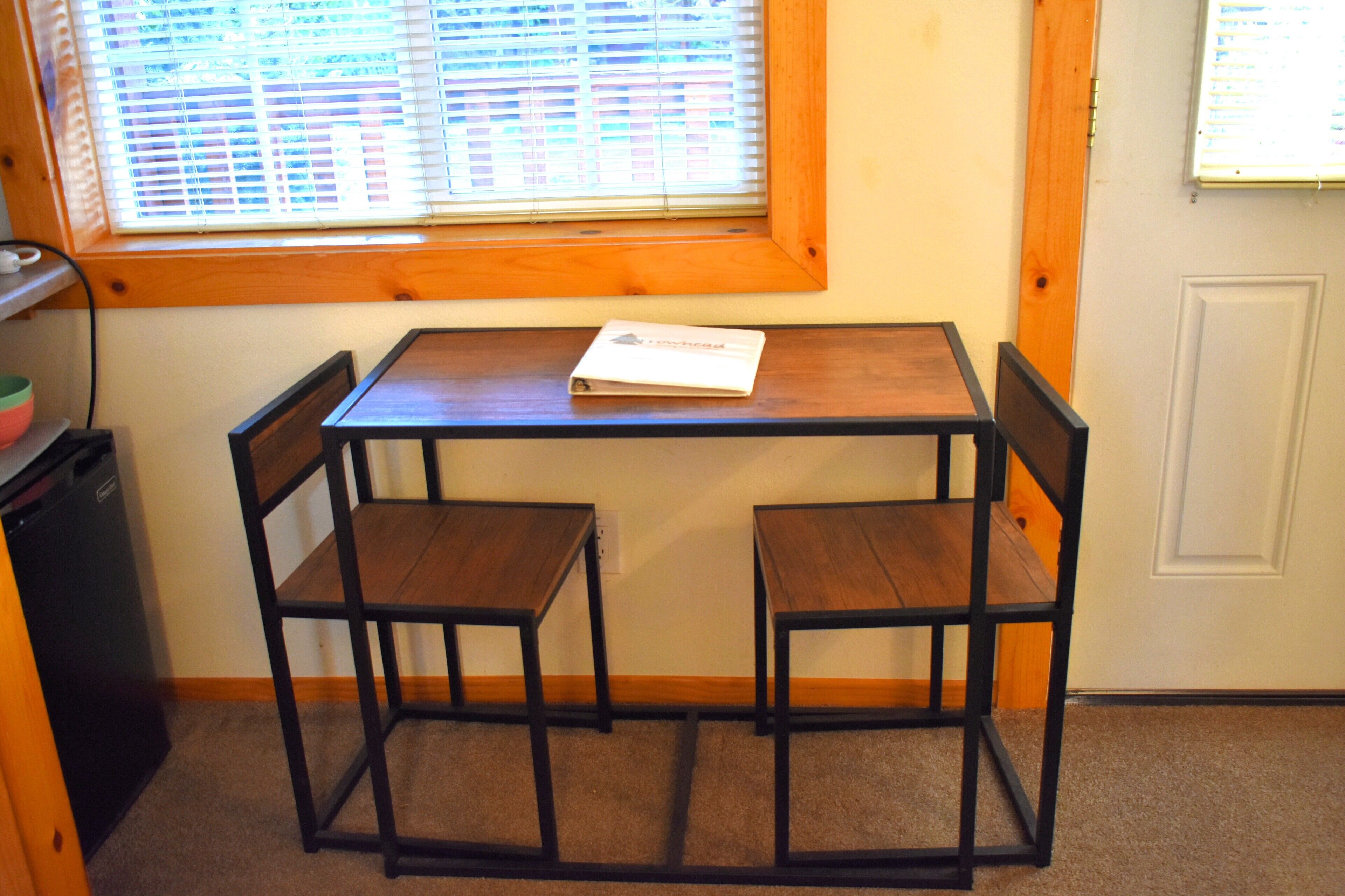 Une petite pièce avec une table en bois et deux chaises, une fenêtre avec des stores et une porte.