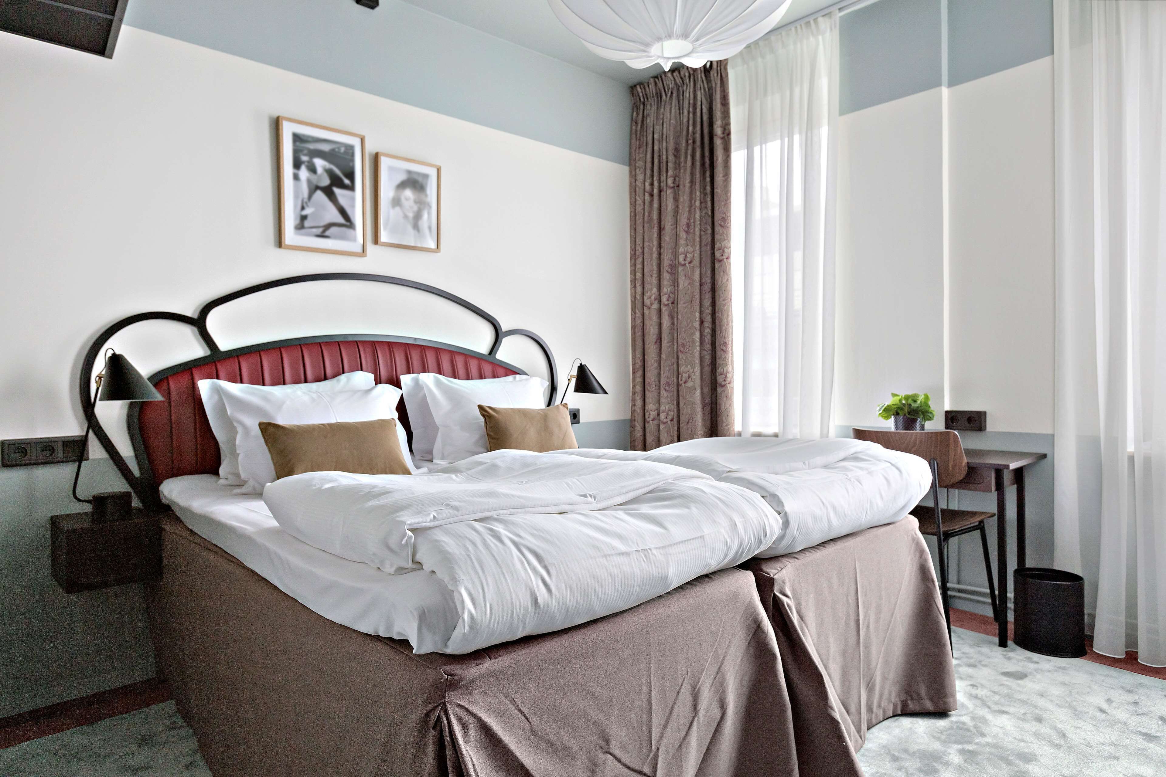 A neatly made bed with white linens, a headboard with a red panel, and a bedside table with a lamp. There are two framed pictures on the wall above the bed.