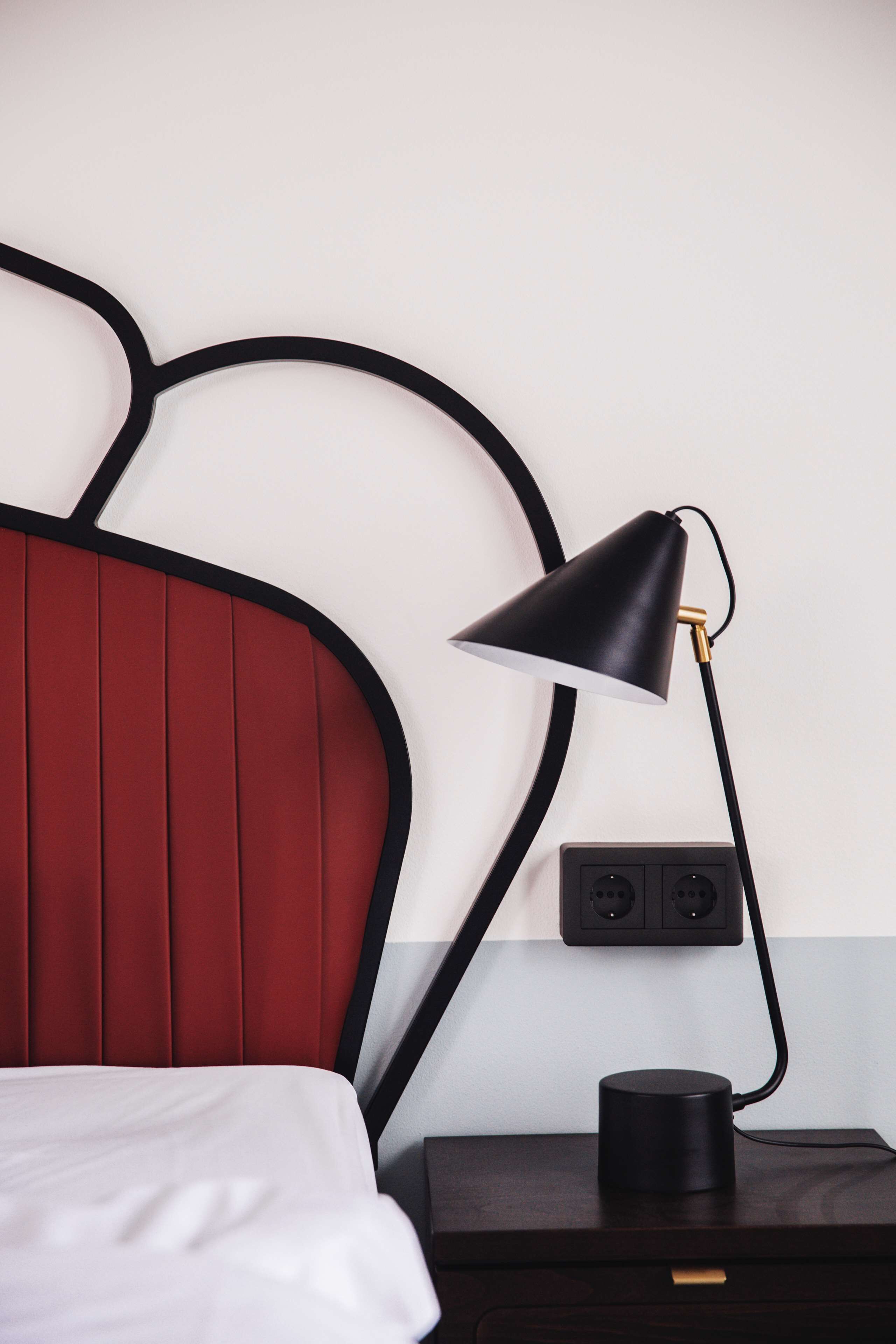 A bed with a red headboard, a black desk lamp, and a black power strip on a wooden nightstand.