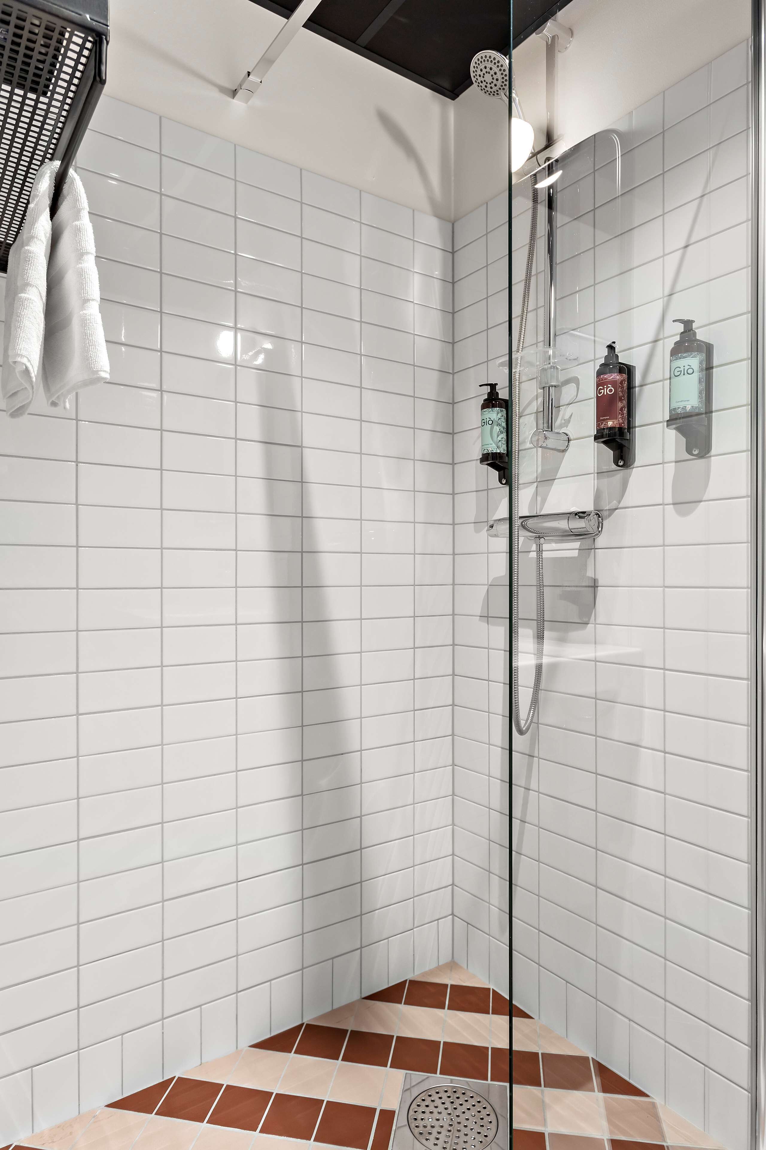 Un área de ducha moderna con mampara de vidrio, paredes cubiertas de azulejos blancos y piso de baldosas marrones.