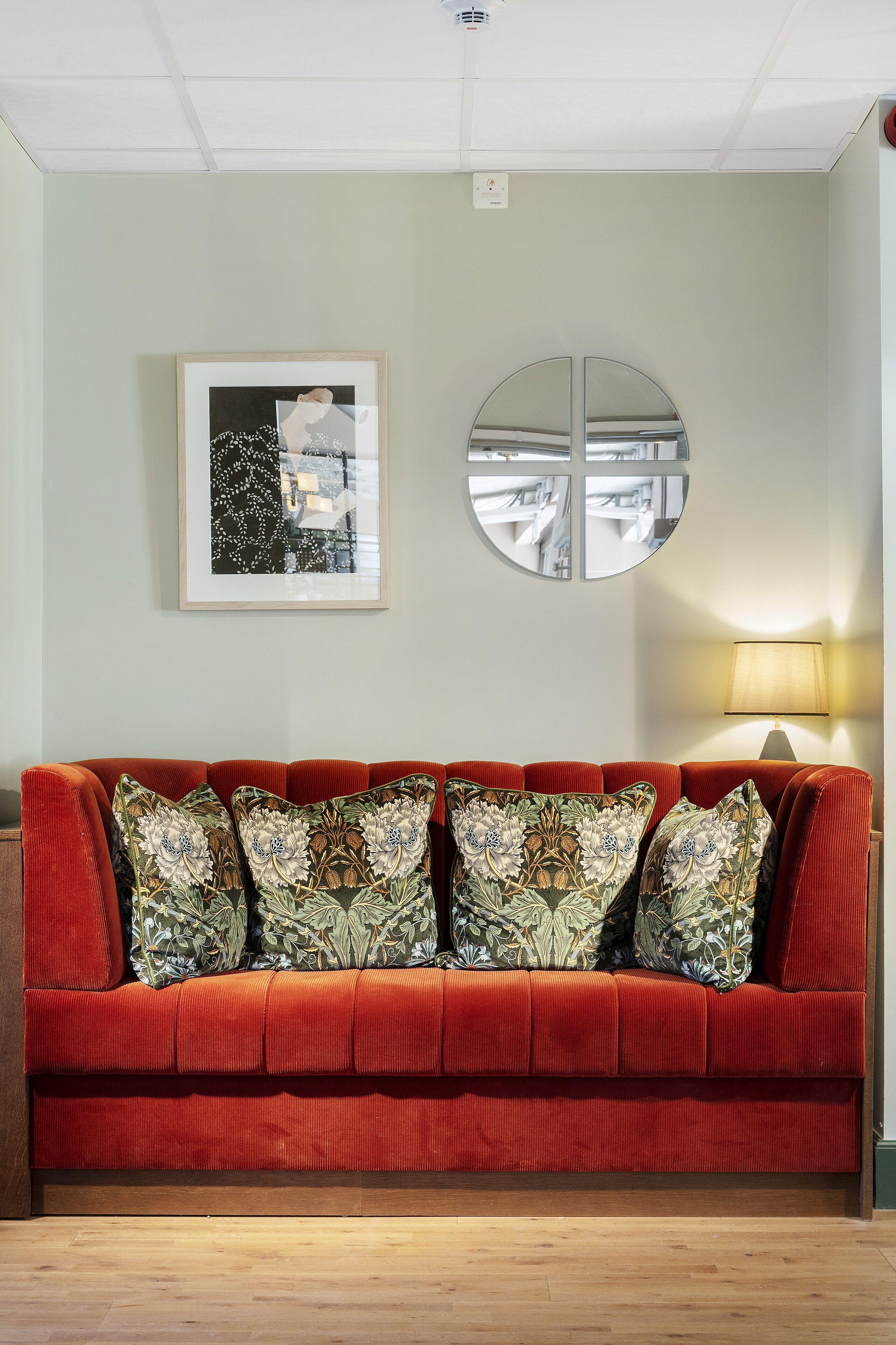A red sofa with floral cushions, a framed abstract painting, and a round mirror on a light green wall.