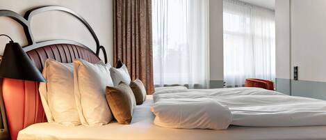 A modern hotel room with a large bed, a bedside lamp, a nightstand, and a view of the city through large windows.