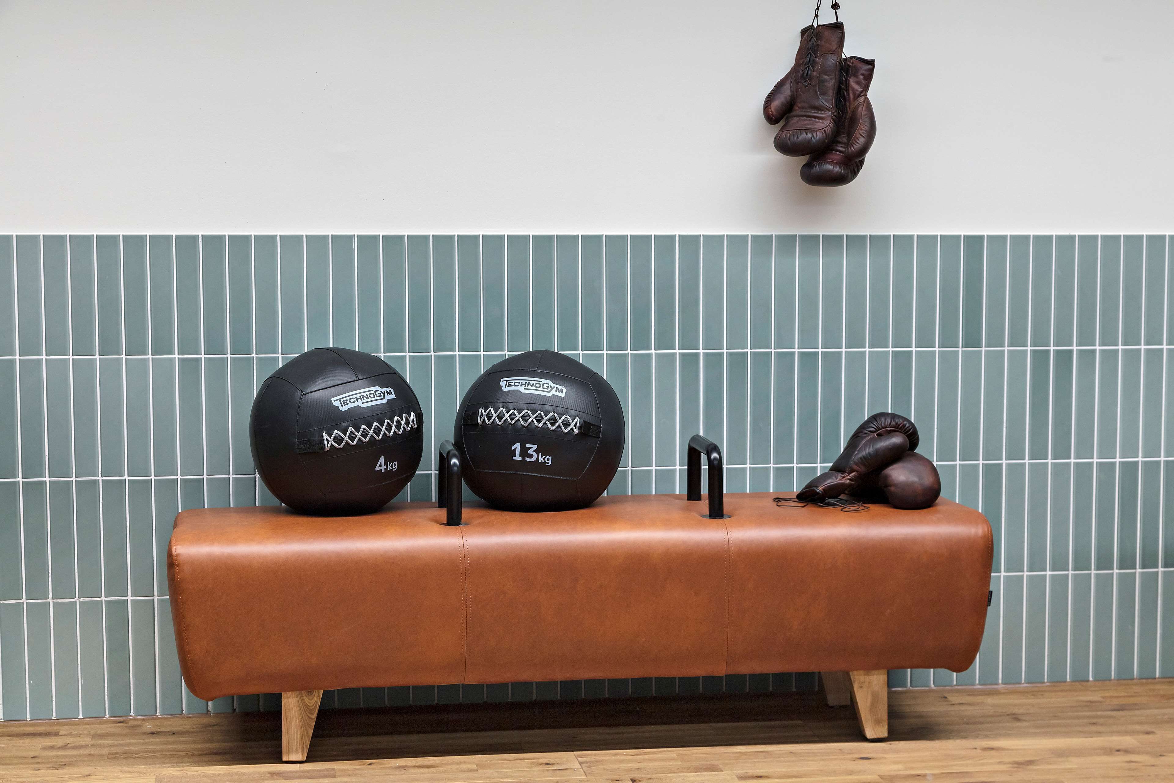 Um banco de couro marrom com duas bolas de exercício pretas e um par de sapatos marrons em frente a uma parede revestida de azulejos.