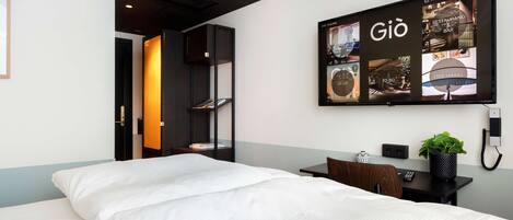 A hotel room with a bed, a desk, a chair, a potted plant, and a wall-mounted television displaying the hotel's name and amenities.