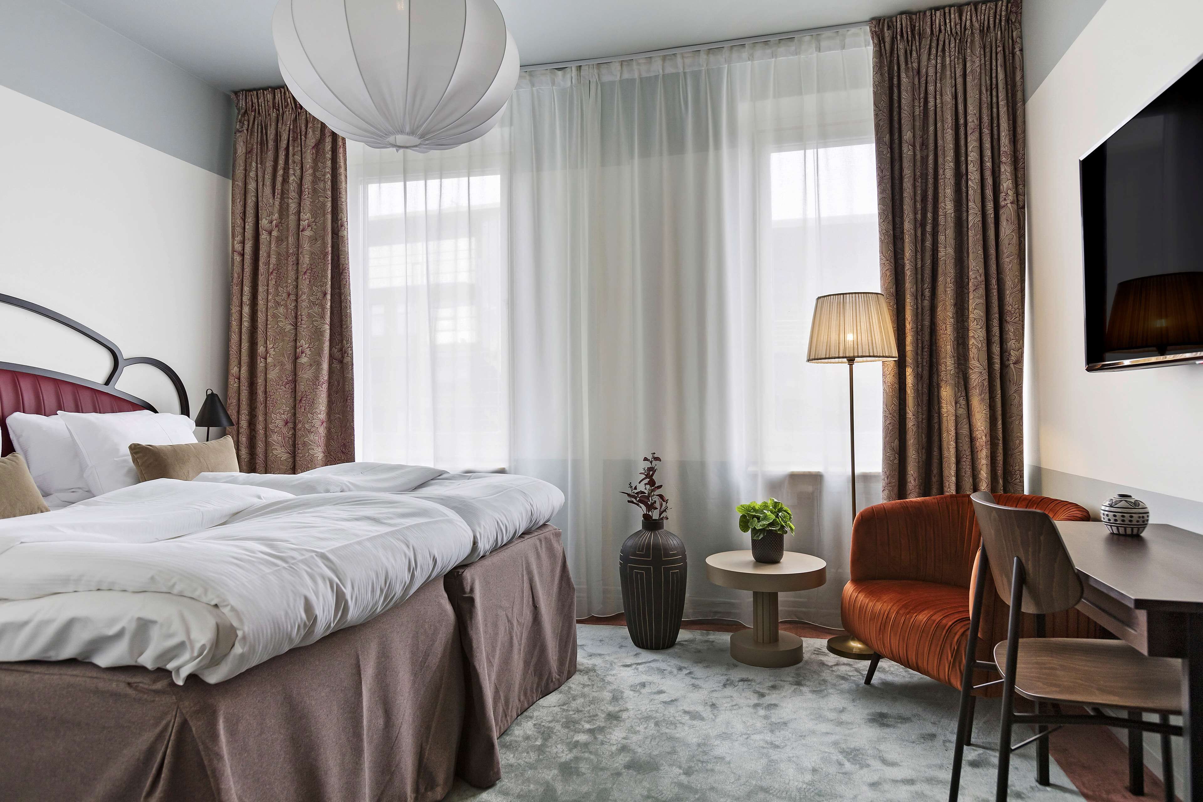 Una habitación de hotel con una cama grande, un escritorio, una silla, una lámpara y un jarrón con una planta.