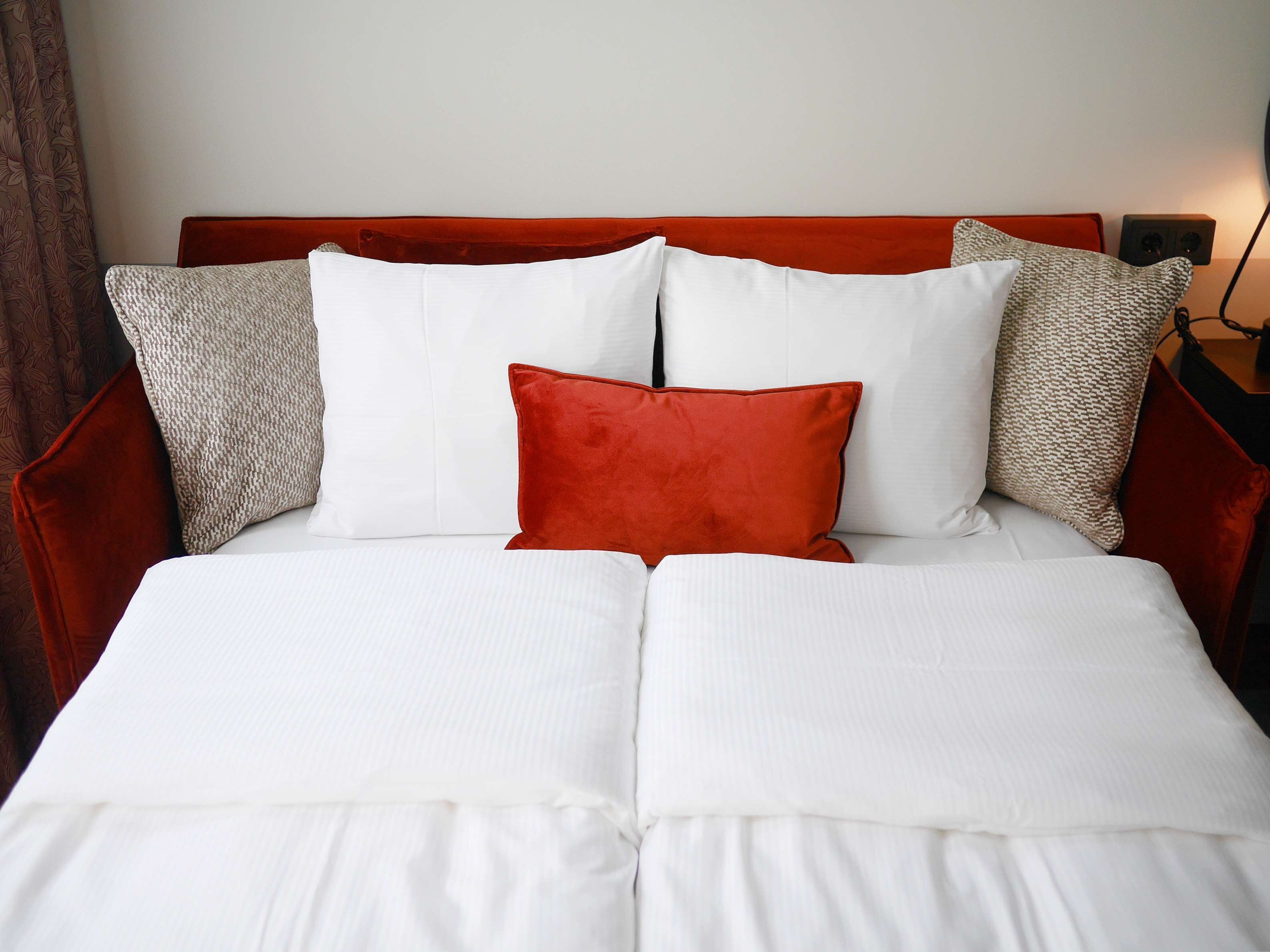 A neatly made bed with white bedding and a red accent pillow, flanked by two patterned pillows.